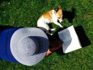 Donna con cappello che legge un libro sull'erba accanto al suo cane di piccola taglia in un giardino soleggiato.