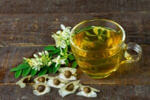 Semi di Moringa marroni e rotondi con ali, disposti su una superficie, con a lato una tazzina di tè e foglie e fiori dell'albero miracoloso.